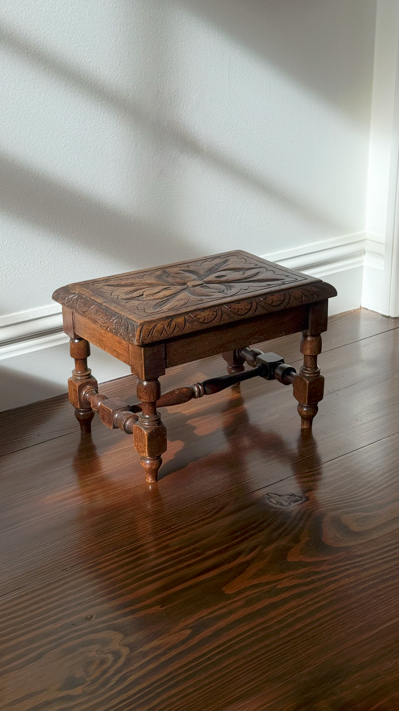 Wooden stool with intricate carvings on a wooden floor against a white wall.